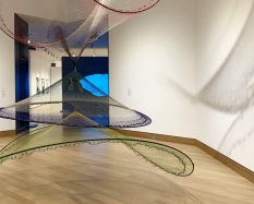 A photograph of an exhibition installation which includes a an Australian Aboriginal painting and a large netted sculpture that hangs from the ceiling. 