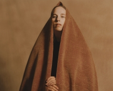 A photograph of a woman looking down into the camera lens. She is wrapped in a tan blanket from head to toe. You see her hands and face. The background is a lighter tan then the blanket that surrounds the subject.