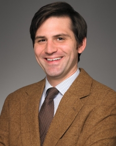 A white man in his 40s stands in quarter profile but faces the camera smiling. He is wearing a brown suit jacket, has brown eyes, and brown hair.