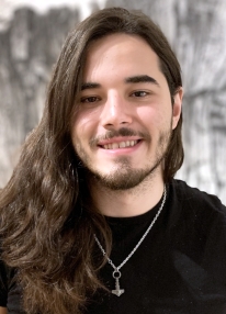 A man in his early 20s with long dark hair, dark eyes is photographed from the chest up. He is wearing a black t-shirt.