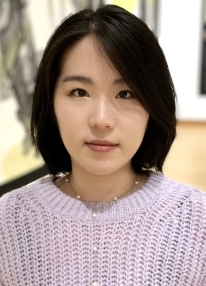 A young women with dark hair, brown eyes, and fair skin is photographed from her chest up. She is wearing a lilac sweater.