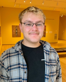 A young man with fair skin, blonde hair, brown eyes, and glasses smiles at the camera. He is photographed from the chest up.