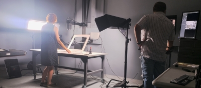 A man and a woman are in a photography room. The woman is working with a two-dimensional artwork on the table in front of her, while the man prepares a computer program and camera to take a photo of the work of art.