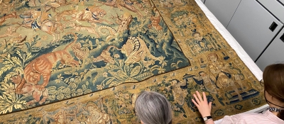 A bird's eye view of a sixteenth-century Flemish tapestry featuring wild animals such as tigers in a forest or jungle setting. The top of two women's heads are visible at the bottom of the photo.