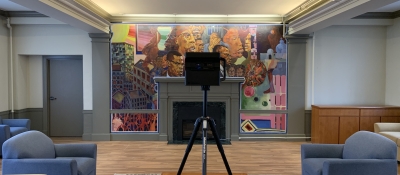 A 3D camera stands on a tripod in a dorm common room. The walls are covered in a fresco mural.