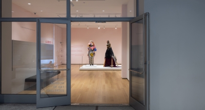 The photographer is standing outside of a museum gallery with two glass doors and a partial glass wall. One of the doors is opening into a brightly lit room painted pink with two larger-than life figurative sculptures using found materials.