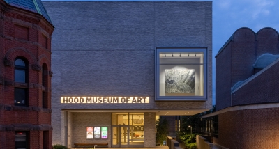 The facade of the Hood Museum of Art at dusk. The building is made of gray bricks and the facade has a large vitrine window on the second floor displaying a black and white work carved into aluminum. The work gives the illusion of moving fabric.