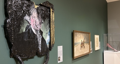 A museum gallery installation with gray and forest green walls. In the foreground of the installation image is a work of art that looks like a portrait of President Washington in a book that has been burnt.