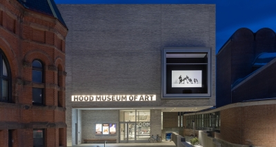 A photograph of the Hood Museum of Art's north facade and entrance taken after sunset. There is a window on the facade displaying a black and white video work.