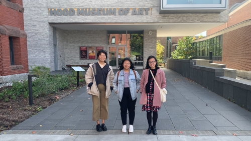 Three women stand outside of the Hood Museum of Art's white bricked facade. They are smiling for the camera. It is a sunny day with blue skies.