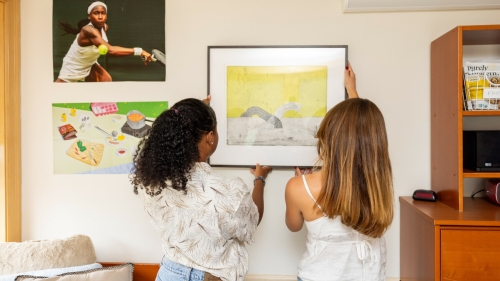 Two students stand facing the wall. They are hanging a painting on the wall of their dorm room. 