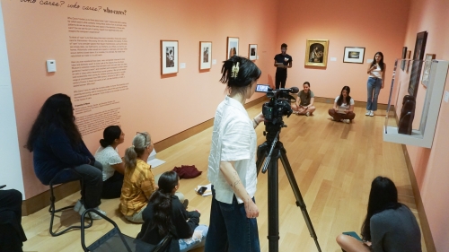 A group of college students film a class discussion in a museum gallery.