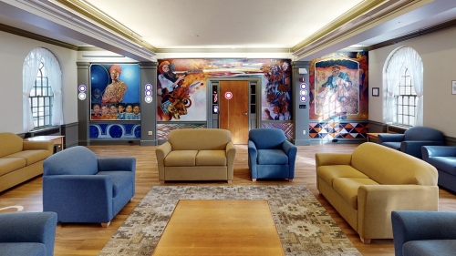 A dormitory common room with blue chairs and tan couches in the center, surrounded by colorful murals.