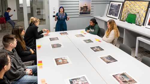 Students sit around a large table, looking at art in one of the BCOS classrooms. A professor stands at the far end of the table, instructing the class.