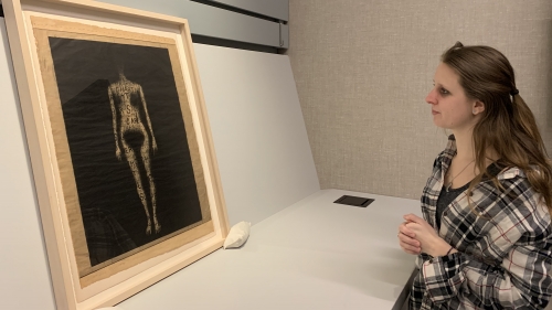 A young woman looking at a framed work of art on paper, that depicts a nude woman, without any features, on a black background.