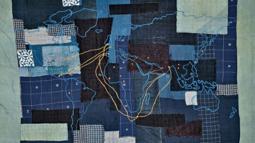 A blue patchwork quilt, with a map of the world embroidered in blue thread. Lines connecting countries are stitched in gold and green thread, and locations on the map are dotted with red thread.