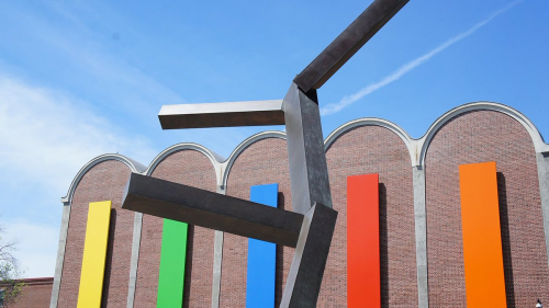 A tall bronze sculpture made up of 5 rectangular prisms which extend like arms and legs from a central "torso". Behind this sculpture is a brick wall made up of 5 arched bays containing rectangular panels in yellow, green, blue, red, and orange.