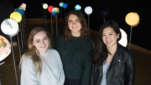 three female students standing together among an art installation of lit up globes