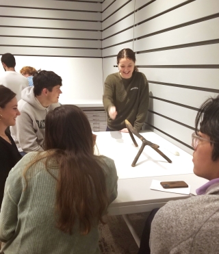 A group of college-age students sit around a white rectangular table and examine an object made of wood.