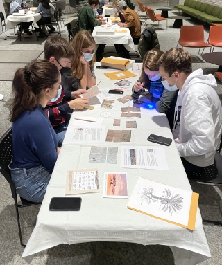 College-age students sit around a rectangular table in the middle of a large atrium space. The students are solving puzzles together on the table in front of them.