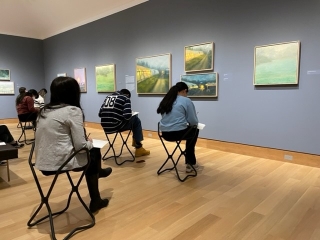 A group of college-aged students sit on stools in a museum gallery in front of oil paintings mostly of abstracted or slightly abstracted landscapes. They are all writing with pencils in notebooks.