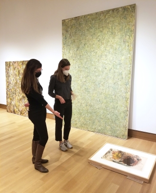 Two women, wearing medical face masks, stand in a museum gallery in which paintings are leaning against the wall in preparation to hang them on the walls. The two women are discussing a work on the floor and the layout of the installation overall.