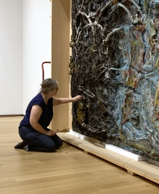 A woman is sitting on a wood floor in a museum gallery. She is working on cleaning a enormous work of art that is an assemblage of found objects, cloth, and paint.