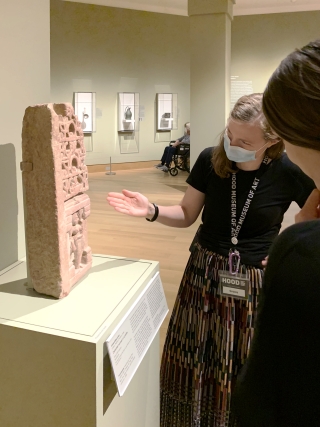 A woman wearing a light blue medical face mask is pointing at an ancient sculpture that appears to be mainly text and talking about it with someone else. Only the back of the second person's head is visible.