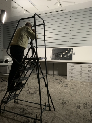 A photographer stands on a ladder while photographing a work of art in a dark classroom.