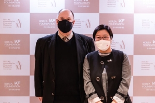 A man and a woman stand side by side linking arms in front of a backdrop printed with a repeated red logo. They are both wearing medical face masks.