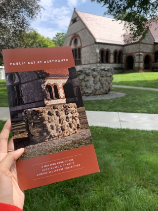 A woman's hand is holding a brochure about public art sculpture on a college campus. In the background of the photo is the work of art and surrounding scenery that is featured on the cover of the brochure.