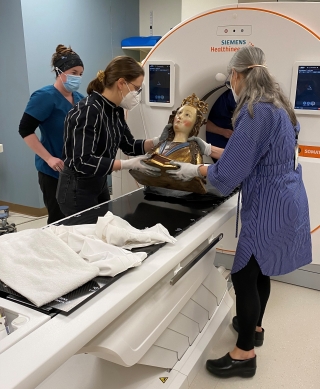 Two women hold an ancient reliquary bust of a female saint and are carefully placing it on an MRI machine to be scanned.