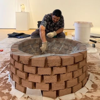 At the center of the photo, a man crouches over a circular clay brick fire pit sculpture. The bricks are a light brown, tinged with red, and there is netting spread over the top. The man, an artist, is putting the final touches on his sculpture.