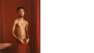 A young man with medium-dark skin tone stands in a red room and wears a red towel around his waist but no shirt. The sitter's expression is serene and he stares at the camera.