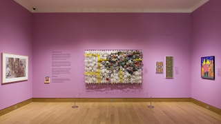 A museum gallery with lilac purple walls and colorful works of art hanging on the walls. There's a large work in the center of the installation made of primarily made of paper and wire.