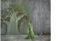 A dark skinned woman wrapped in a green robe stands to the right of a tree painted on a grey wall. 