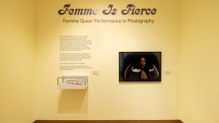 A photograph of a gallery with yellow walls. In purple glitter vinyl, above a photograph of the Black person in a fur coat, is says "Femme is Fierce."