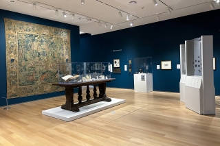At the center of a room with blue walls sits a large wooden table with a glass case containing multiple sculptures of various sizes, shapes and materials. Behind it, hangs a large colorful tapestry. 