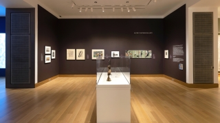 A museum gallery installation with works hanging on a dark purple wall and a case in the middle of the gallery with sculpture in it.
