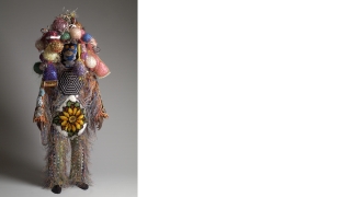 A life size sculpted human-like figure is covered in colorful knitted fabrics, plastic netting, Easter grass, sequins, mirrors, beads, yarn, and paint.