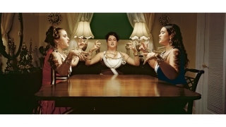 A photograph of three women sitting around a table. They appear to be preforming or magic of some kind. Their eyes are closed and mouths are open as if they are chanting. Their beaded necklaces float in the air, off of their necks.