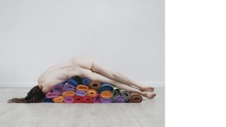 A nude woman lays on her side on top of a stack of rolled, colorful yoga mats. Her back is to the camera.