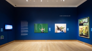 A museum gallery painted with dark royal blue walls. There are three large framed photographs and a television displaying digital artwork as well.