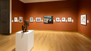 A photograph of a museum gallery installation. The walls are a deep orange-brown. There are framed works hanging on the walls.