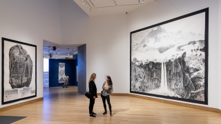 Two women stand in a museum gallery surrounded by gigantic works on paper of mountain landscapes.