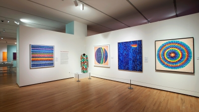 A photograph of a museum gallery installed with the work of female artist Alma W. Thomas. Her works are bright and colorful, they are hung on a white wall. There is also clothing the artist made that is also bright, colorful, and textured.