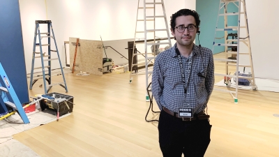 A young man with dark hair and glasses stands in a museum gallery that is being painted. He is facing the camera and smiling, his hands are behind his back. He is wearing a white and blue checkered shirt with black pants.