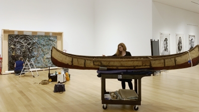A woman stands in a museum gallery with white walls and high ceilings. She is using a brush to clean a Native American birch bark canoe.