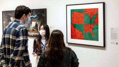 Three college-aged students stand in a museum gallery and look at a map of the USA that divided into quadrants of bright red and green. One of the students face us and is talking to the other two students about the work of art.