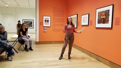 A college-age woman stands in a gallery with bright orange walls and vibrant works on paper hang one them. The woman is talking to the audience and pointing to a work of art in the installation.